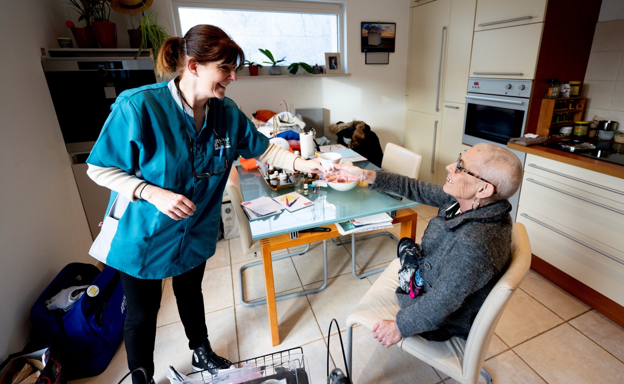 L'Aide Familiale, debout, tend une tasse de café à la Bénéficiaire, qui est assise devant sa table de cuisine. Elles se sourient en se regardant.