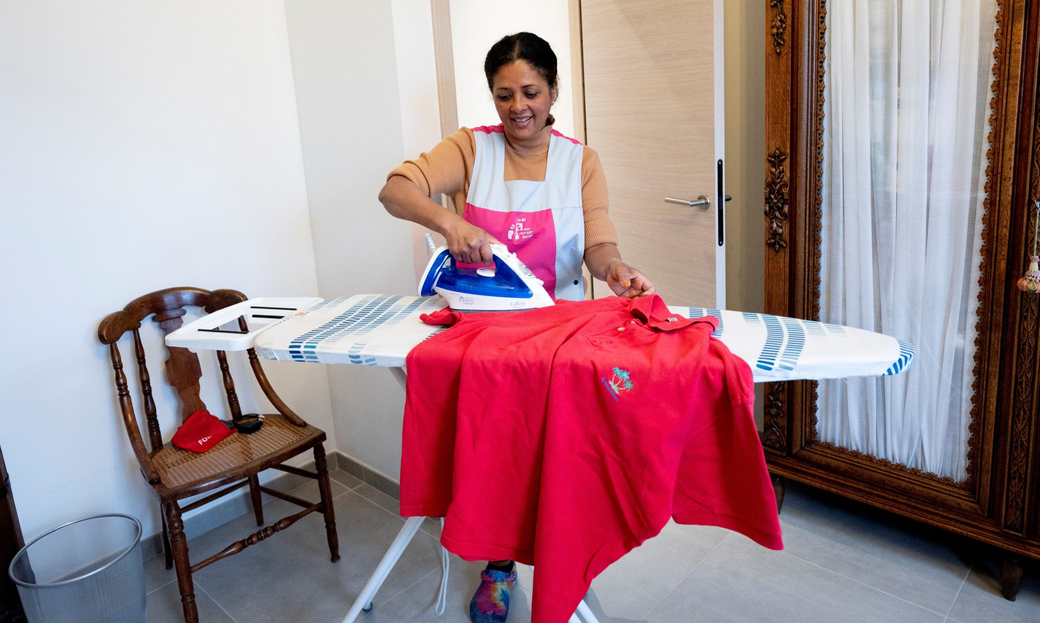 L'Aide Ménagère Sociale, souriante, repasse un polo du Bénéficiaire.