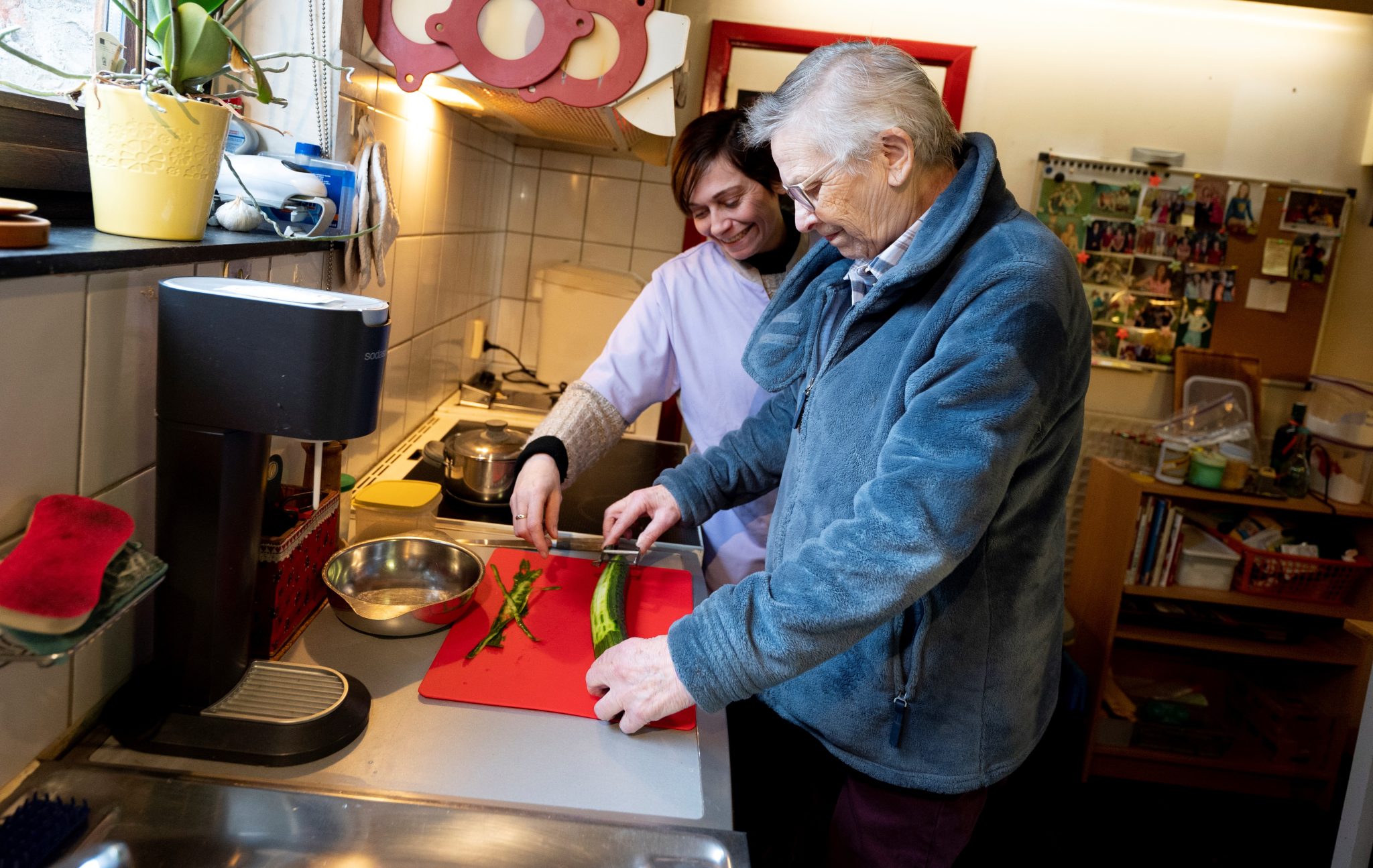 La Bénéficiaire, debout devant le plan de travail de sa cuisine, épluche un concombre. La Garde à domicile Alzheimer, à ses côtés, la guide.