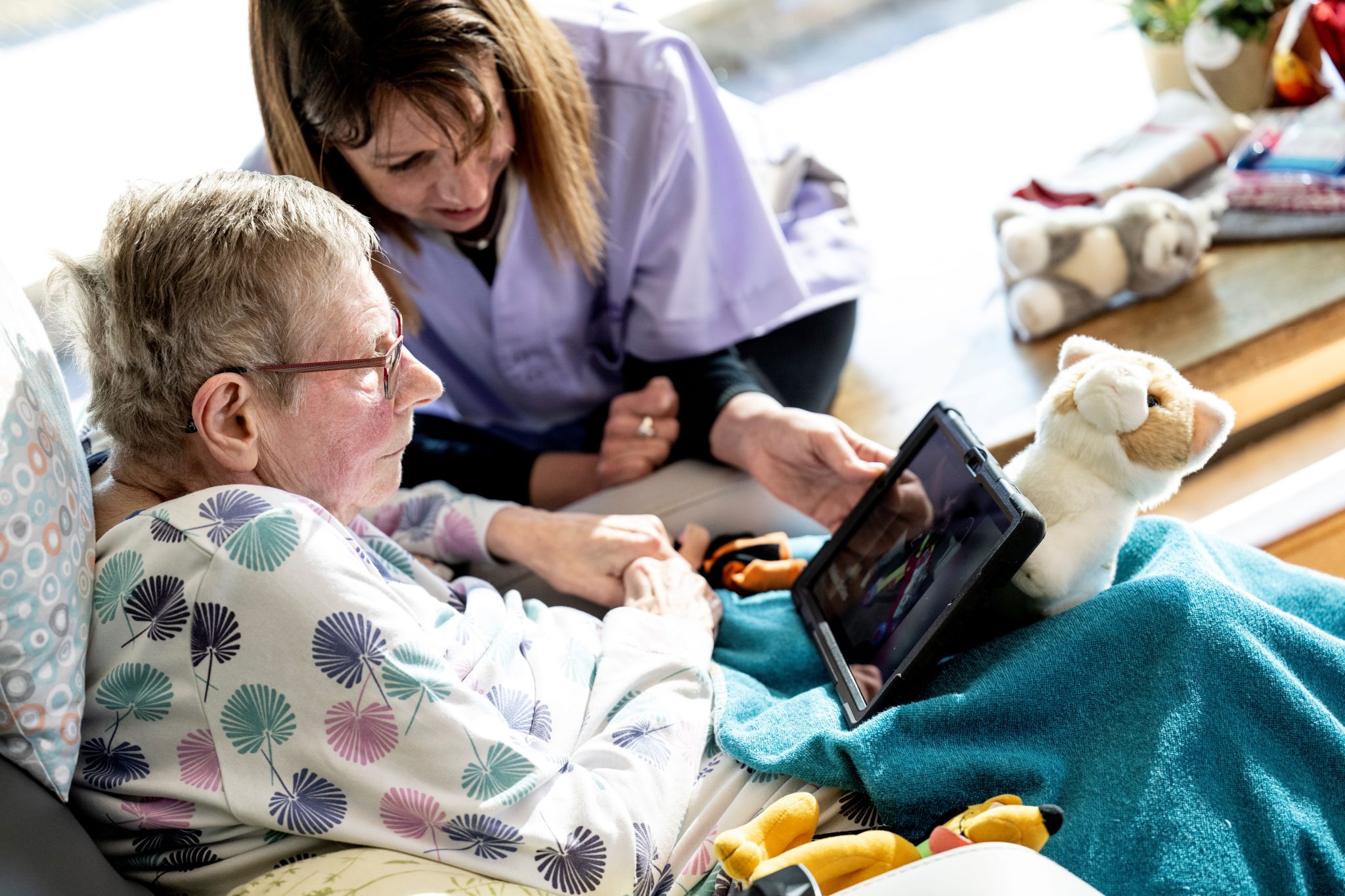 La Garde à domicile, aux côtés de la Bénéficiaire, tient la tablette numérique interactive sur les genoux de la Bénéficiaire, qui est installée dans son fauteuil. Elles regardent des vidéos sur l'écran.