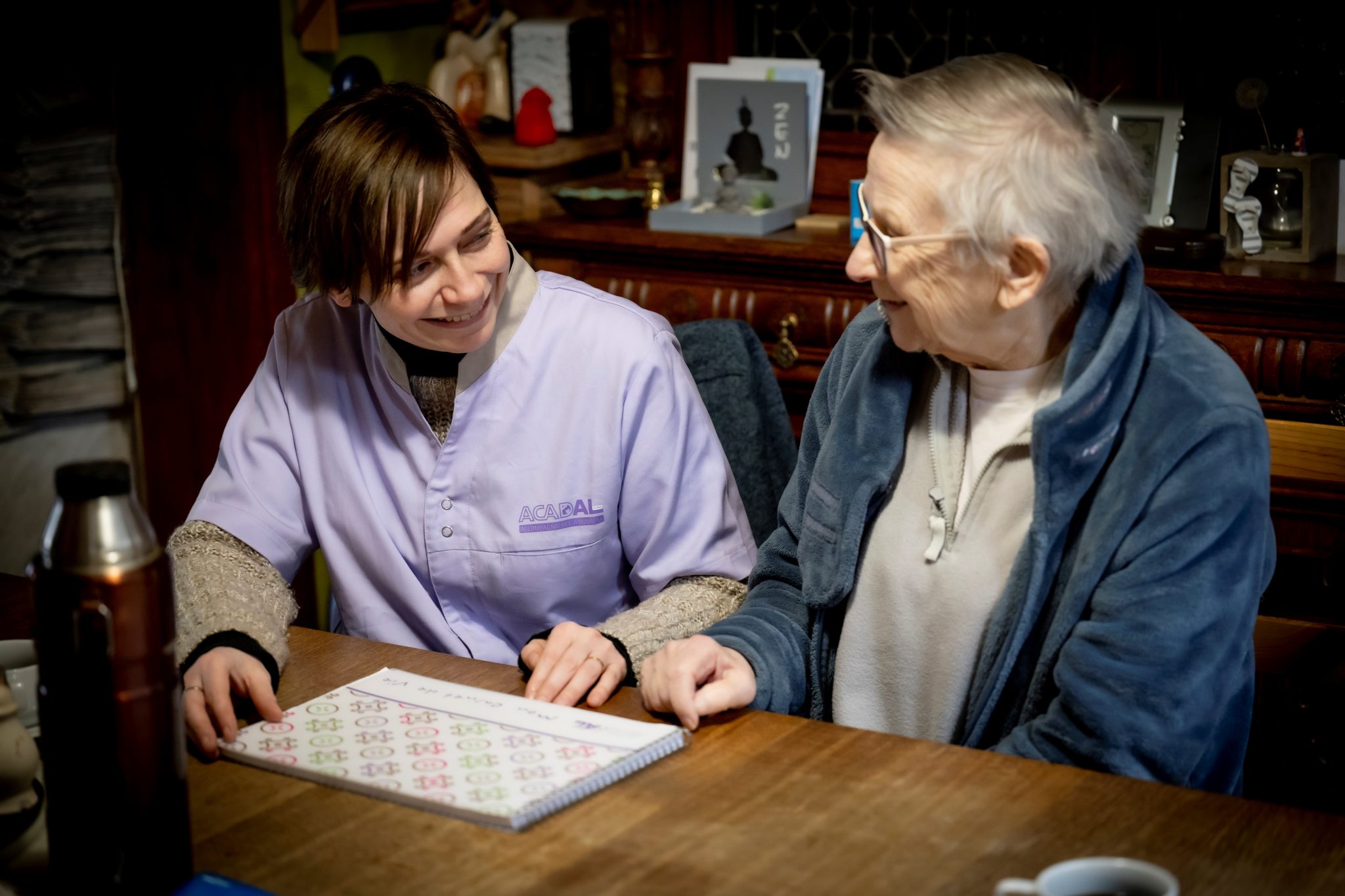 La Garde à domicile Alzheimer et la Bénéficiaire sont attablées et s'apprêtent à ouvrir le Carnet de Vie pour le feuilleter.